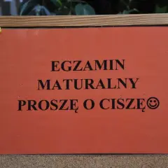 Pomarańczowa kartka na tablicy korkowej z napisem "EGZAMIN MATURALNY PROSZĘ O CISZĘ" i uśmiechniętą buźką.