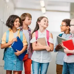Grupa uśmiechniętych dzieci z plecakami i książkami stoi w szkolnym korytarzu.