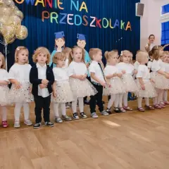 Dzieci w strojach galowych stoją w rzędzie na tle niebieskiej kurtyny z napisem "Pasowanie na przedszkolaka".