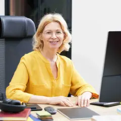 Uśmiechnięta kobieta w okularach i żółtej bluzce siedzi przy biurku z komputerem.