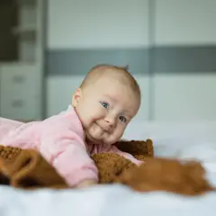 Uśmiechnięte niemowlę w różowym pajacyku leży na łóżku obok brązowego kocyka.