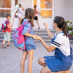 Kobieta klęczy przed dziewczynką z plecakiem, rozmawiając z nią przed szkołą.