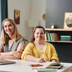 Dwie kobiety siedzą przy biurku, jedna z zespołem Downa, obie uśmiechnięte.