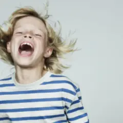 Chłopiec z blond włosami śmieje się głośno, z otwartymi ustami i rozwianymi włosami.