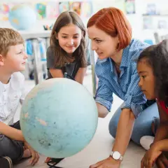 Nauczycielka z rudymi włosami pokazuje dzieciom globus w klasie.