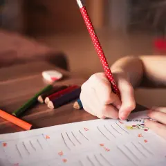 Dziecko pisze kredkami na kartce, obok leżą kolorowe kredki i gumka.