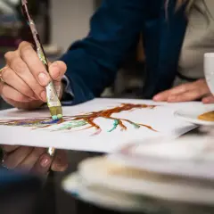 Dłoń maluje pędzlem na białej kartce, obok filiżanka kawy i ciasteczko.