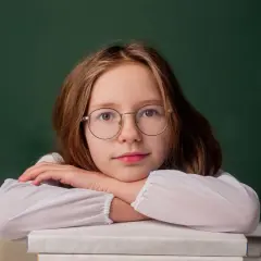 Dziewczynka w okularach opiera głowę na stosie książek przed zieloną tablicą.