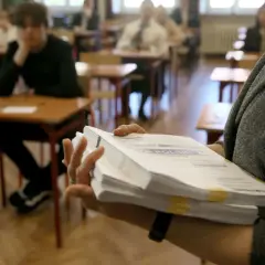 Ręka trzyma stos arkuszy egzaminacyjnych z napisem "JĘZYK POLSKI". W tle uczniowie siedzą przy ławkach.