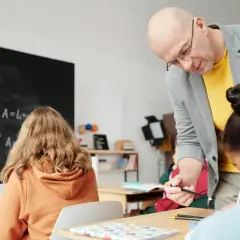 Nauczyciel pomaga uczniom przy biurkach w klasie, na tablicy widać wzory matematyczne.
