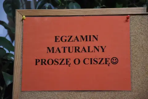 Pomarańczowa kartka na tablicy korkowej z napisem "EGZAMIN MATURALNY PROSZĘ O CISZĘ" i uśmiechniętą buźką.