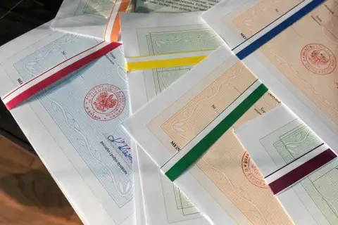 Stos papierów z kolorowymi wstążkami, każdy z pieczęcią i podpisem dyrektora. To świadectwo ukończenia szkoły.
