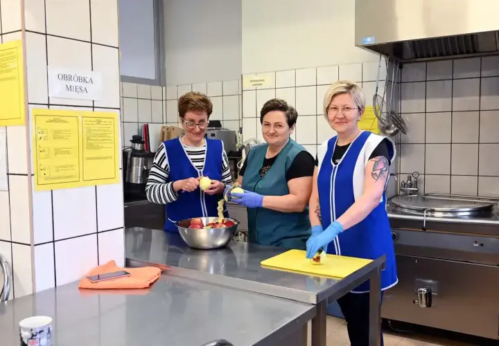 Trzy kobiety w fartuchach obierają jabłka w szkolnej kuchni.