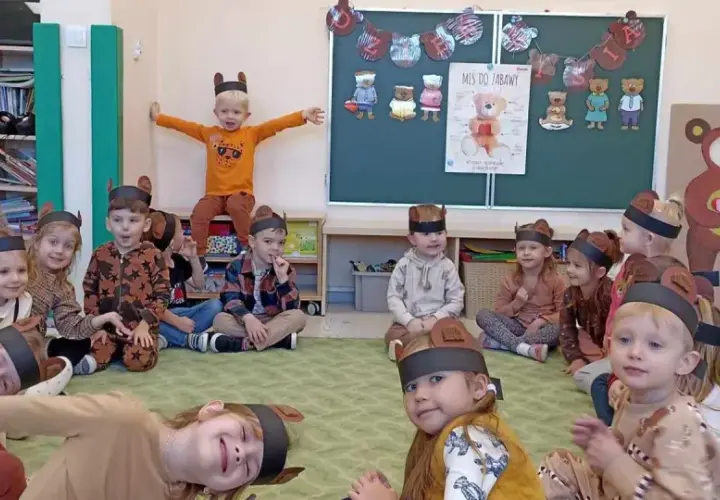 Dzieci w strojach misiów siedzą na dywanie w sali przedszkolnej.