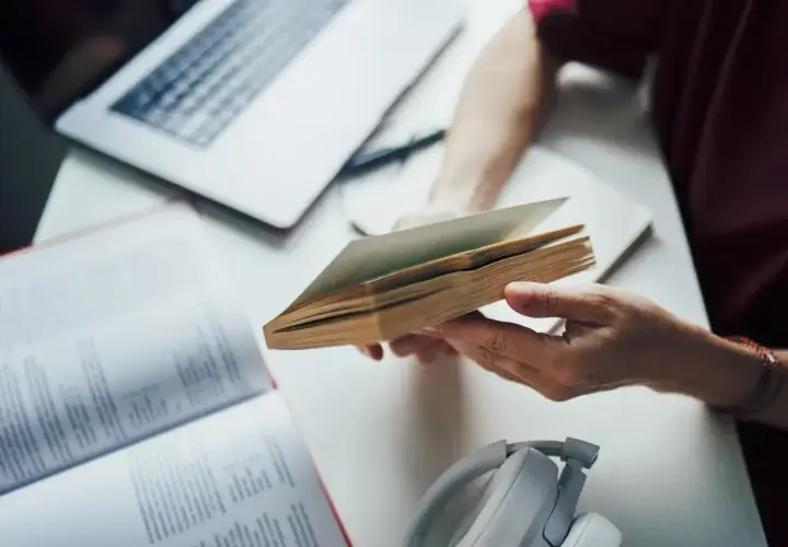 Osoba przegląda książkę, przygotowując się do napisania bibliografii. Obok leży laptop i słuchawki.