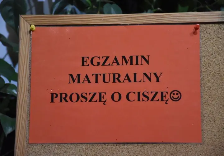 Pomarańczowa kartka na tablicy korkowej z napisem "EGZAMIN MATURALNY PROSZĘ O CISZĘ" i uśmiechniętą buźką.