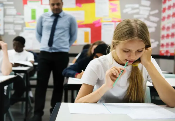 Dziewczynka z ołówkiem w ustach pisze test, nauczyciel stoi z tyłu.