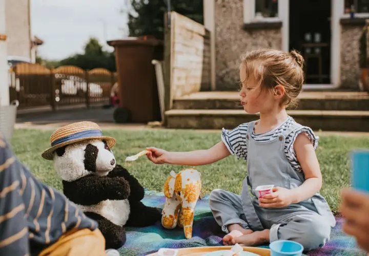 Dziewczynka karmi pluszową pandę w kapeluszu na pikniku na trawie.