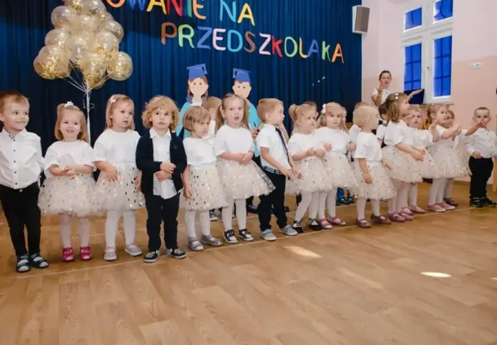 Dzieci w strojach galowych stoją w rzędzie na tle niebieskiej kurtyny z napisem "Pasowanie na przedszkolaka".