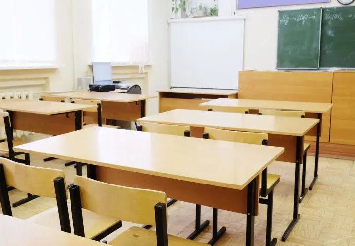 Pusta klasa szkolna z rzędami drewnianych ławek i krzeseł, tablicą i projektorem.