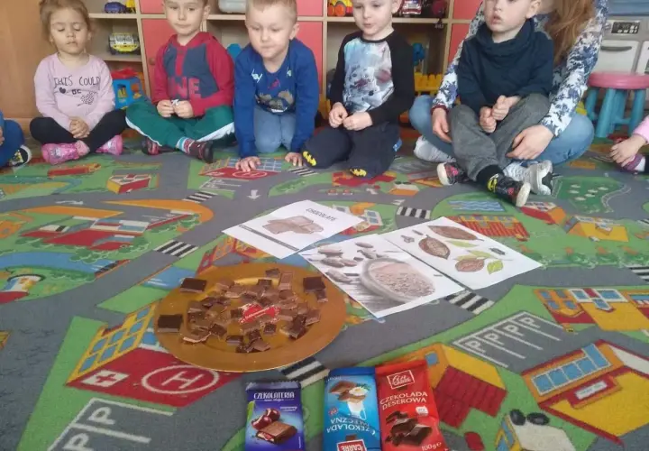 Dzieci w przedszkolu siedzą na dywanie z zabawkami, przed nimi talerz z kawałkami czekolady i opakowania czekolad.