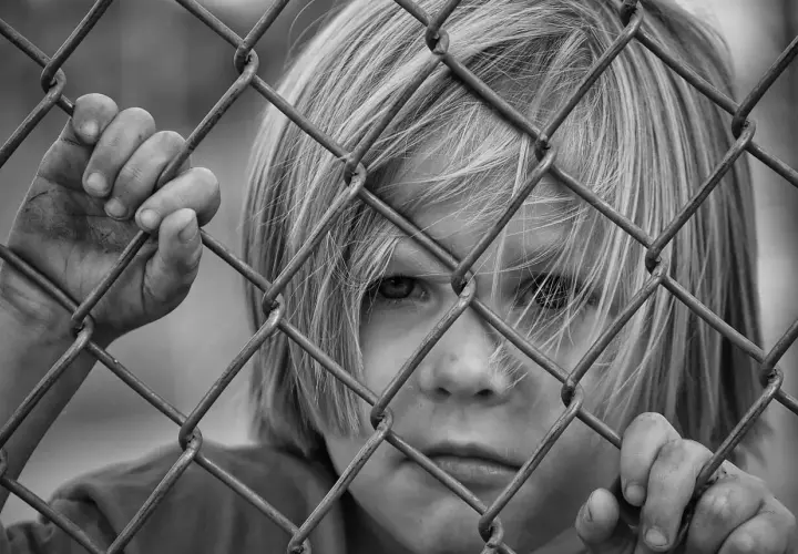 Czarno-białe zdjęcie dziecka z blond włosami, patrzącego przez siatkę ogrodzeniową.