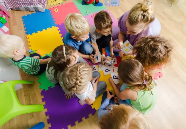 Dzieci i dorosła siedzą na kolorowych puzzlach, grając w karty z literami.