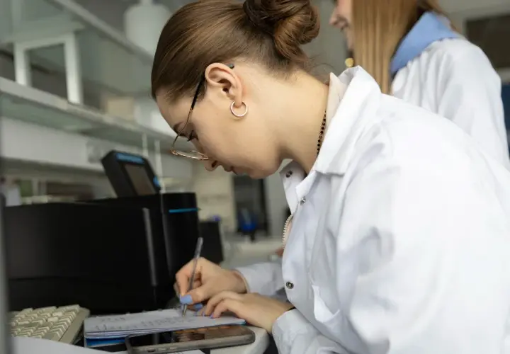 Studentka w białym fartuchu laboratoryjnym pisze notatki w zeszycie, obok klawiatury i telefonu.