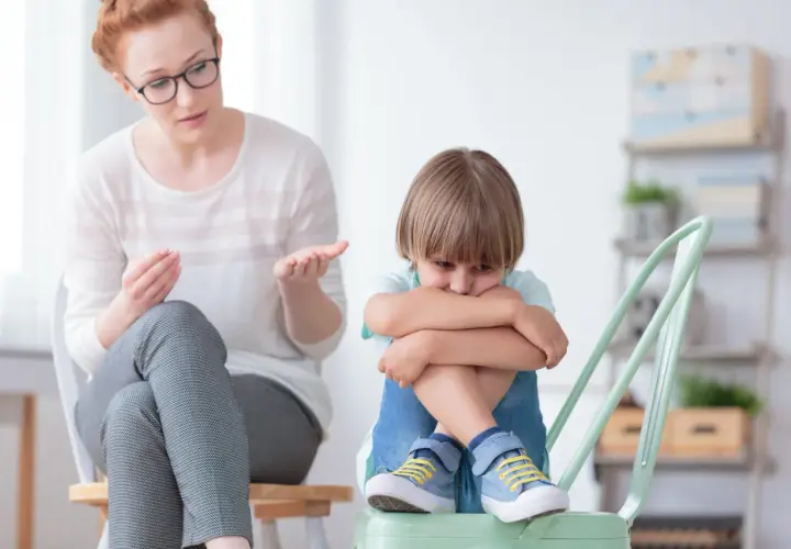 Kobieta w okularach rozmawia ze smutnym chłopcem siedzącym na krześle.