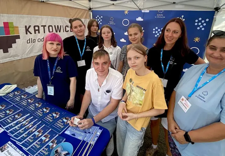 Grupa osób, w tym młodzież i dorosłych, stoi za stołem z książkami i materiałami promocyjnymi "Szkoła w Chmurze".