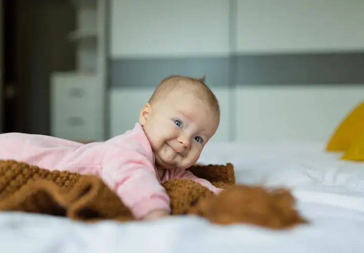Uśmiechnięte niemowlę w różowym pajacyku leży na łóżku obok brązowego kocyka.