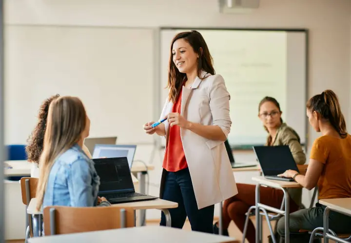 Nauczycielka z markerem prowadzi lekcję dla studentów przy laptopach w sali lekcyjnej.