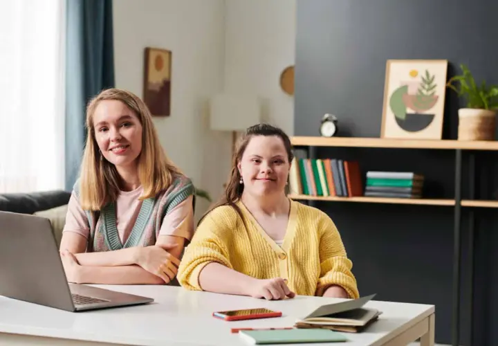 Dwie kobiety siedzą przy biurku, jedna z zespołem Downa, obie uśmiechnięte.