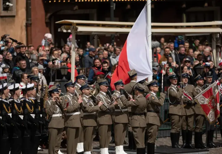 Orkiestra wojskowa grająca na instrumentach dętych na tle tłumu i polskiej flagi.