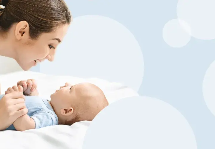 Uśmiechnięta mama pochyla się nad leżącym na plecach niemowlęciem, trzymając je za rączki.