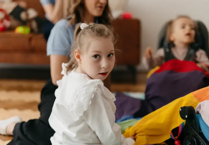 Dziewczynka w białej bluzce patrzy w obiektyw, w tle kobieta i dziecko w wózku.