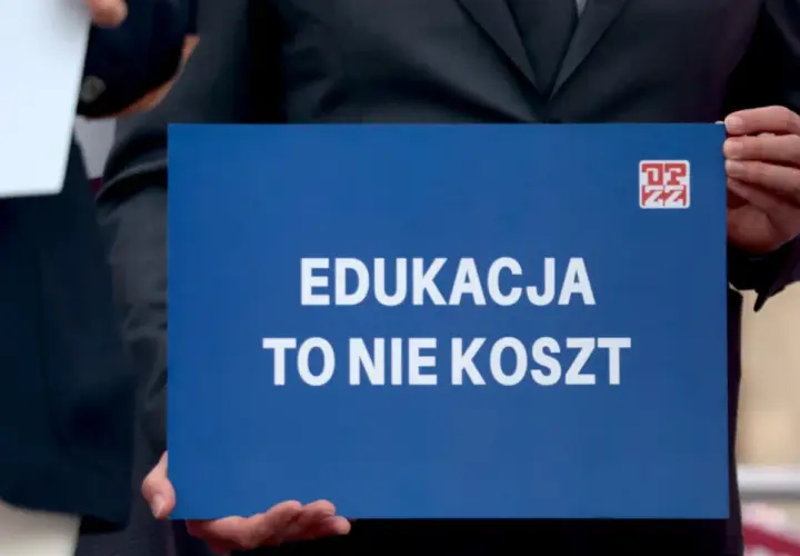 Niebieska tablica z białym napisem "EDUKACJA TO NIE KOSZT" i logo ZZ.