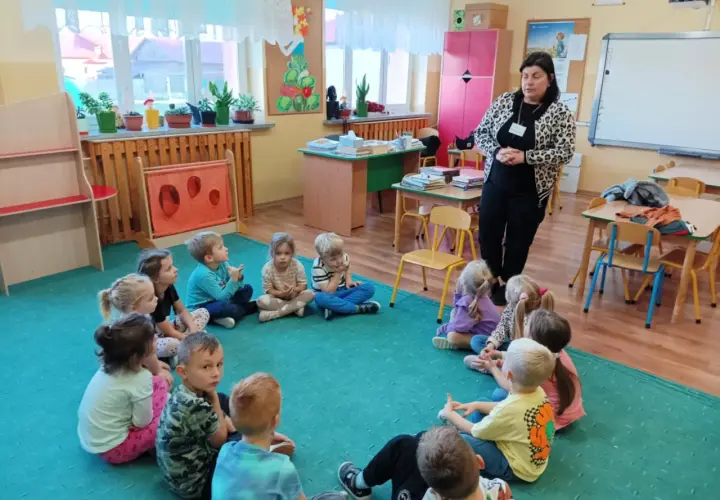Kobieta w panterkowej kurtce stoi przed grupą dzieci siedzących w kręgu na zielonej wykładzinie.
