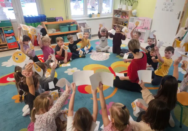 Dzieci w przedszkolu siedzą na dywanie i trzymają w górze rysunki z buźkami.