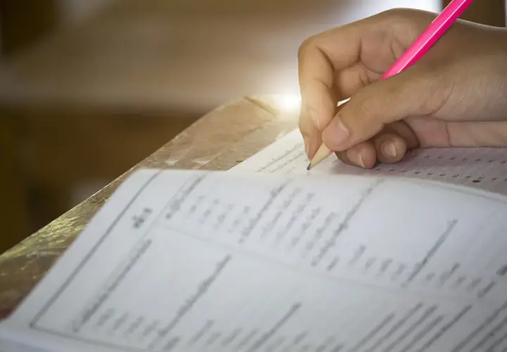 Dłoń ucznia pisząca różowym ołówkiem na arkuszu testowym z kółkami do zaznaczenia odpowiedzi.
