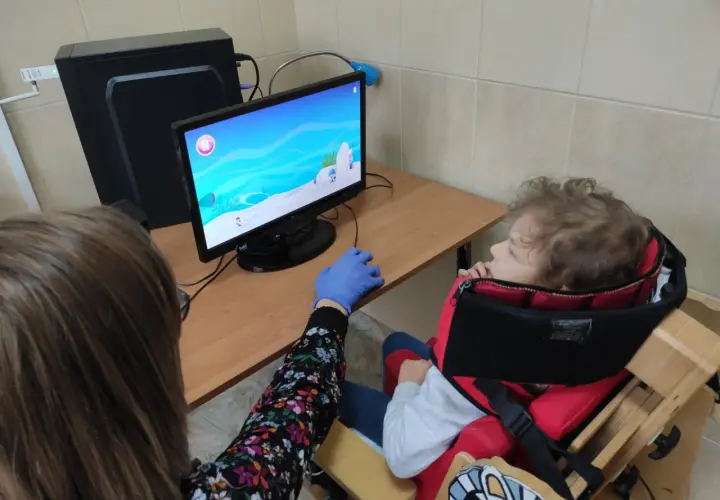 Dziecko w specjalnym foteliku siedzi przed komputerem, na ekranie gra. Obok siedzi osoba w rękawiczce.
