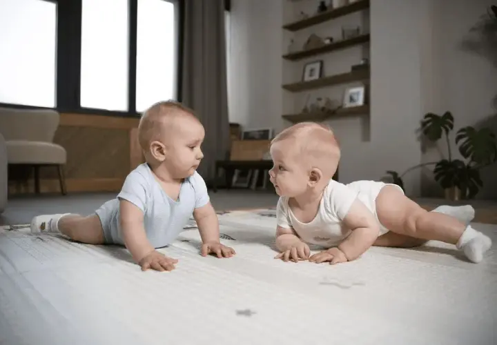 Dwa niemowlęta w body leżą na macie i patrzą na siebie.