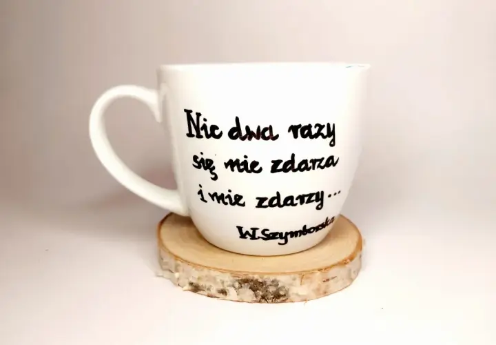 Biały kubek z cytatem Wisławy Szymborskiej "Nic dwa razy się nie zdarza..." na drewnianym podkładzie.