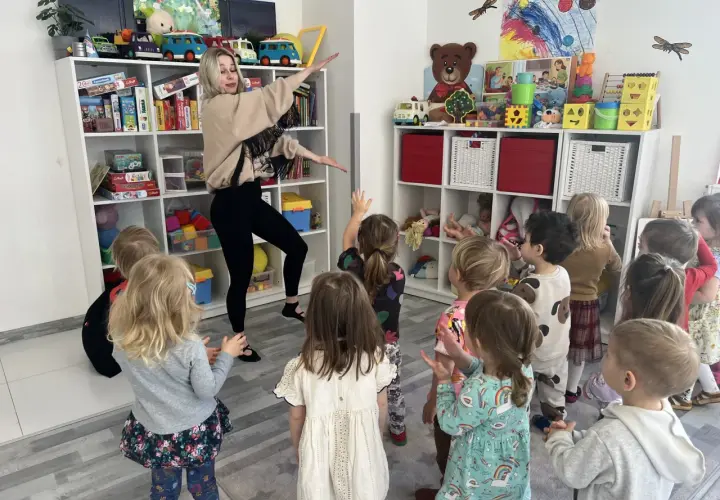 Nauczycielka z przedszkola prowadzi zajęcia rytmiczne dla grupy dzieci, które patrzą na nią z zaciekawieniem.