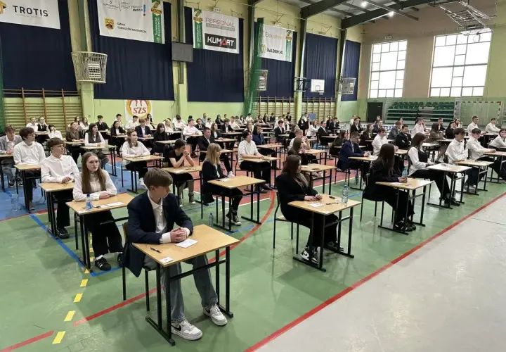 Uczniowie siedzą przy ławkach w sali gimnastycznej podczas egzaminu.