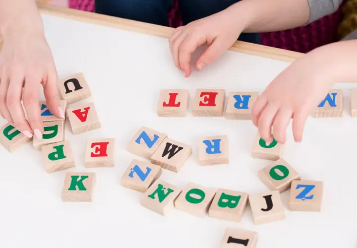 Dzieci układają litery na tablicy, ćwicząc podział na głoski. Widoczne litery to m.in. U, A, T, R, E, N, K, P, Z, O, B, J, I.