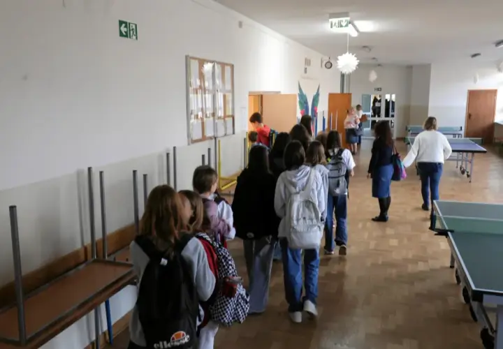 Uczniowie w kolejce na szkolnym korytarzu, w tle stoły do tenisa stołowego.