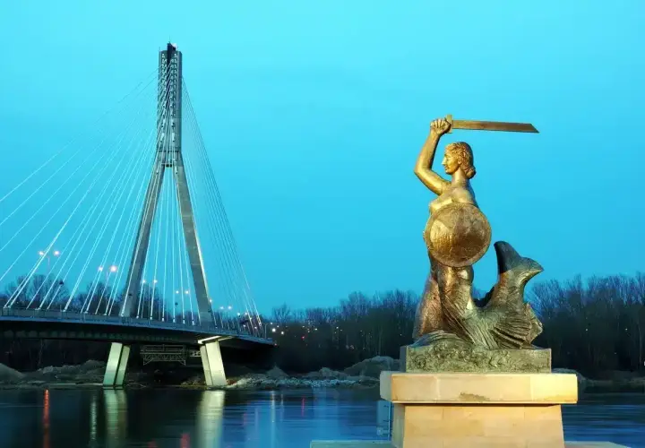 Pomnik Syrenki Warszawskiej z mieczem i tarczą na tle mostu i rzeki.