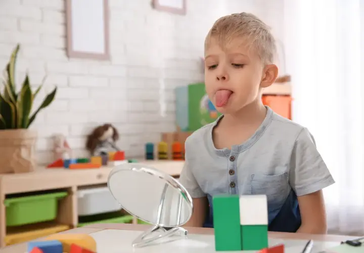 Chłopiec z wysuniętym językiem patrzy w lustro, obok kolorowe klocki.