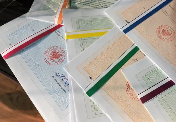 Stos papierów z kolorowymi wstążkami, każdy z pieczęcią i podpisem dyrektora. To świadectwo ukończenia szkoły.